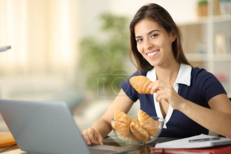 Photo for Happy student e-learning eating croissants looking at camera at home - Royalty Free Image