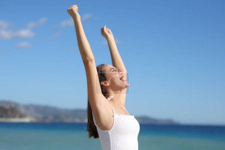 Téléchargez les photos : Femme excitée en robe blanche célébrant lever les bras sur la plage une journée d'été ensoleillée - en image libre de droit