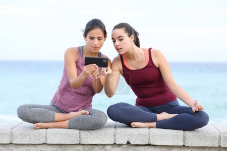 Foto de Retrato vista frontal de dos yoguis atentos viendo tutorial de yoga en el teléfono en la playa - Imagen libre de derechos