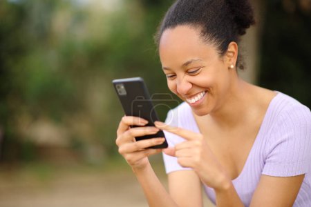 Téléchargez les photos : Heureuse femme noire vérifiant les nouvelles de téléphone intelligent dans un parc - en image libre de droit