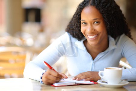 Téléchargez les photos : Heureuse femme noire vous regarde écrire dans l'agenda papier dans une terrasse de restaurant - en image libre de droit