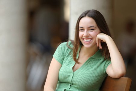 Téléchargez les photos : Femme heureuse riant en regardant la caméra assise sur un banc dans la rue - en image libre de droit
