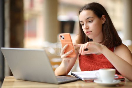 Téléchargez les photos : Femme sérieuse vérifiant téléphone et ordinateur portable dans une terrasse de bar - en image libre de droit