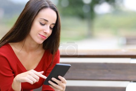Téléchargez les photos : Femme en rouge vérifiant téléphone intelligent assis sur un banc dans un parc - en image libre de droit