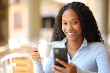 Foto de Mujer negra feliz bebiendo café usando el teléfono en la terraza del restaurante - Imagen libre de derechos