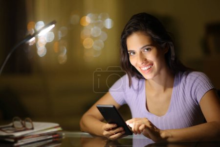 Photo for Happy woman holding phone in the night looks at you at home - Royalty Free Image