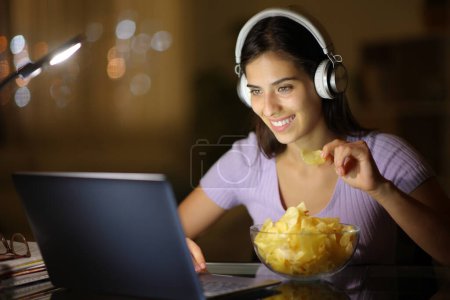 Téléchargez les photos : Femme heureuse portant un casque dans la nuit regarder la vidéo sur ordinateur portable manger des croustilles à la maison - en image libre de droit