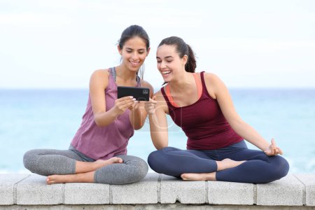 Foto de Vista frontal de dos yoguis felices viendo clases de yoga en línea en el teléfono en la playa - Imagen libre de derechos