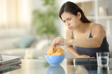 Photo for Glutton woman eating croissants suffering belly ache at home - Royalty Free Image