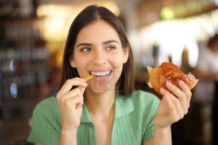 Foto de Mujer feliz comiendo hamburguesa y papas fritas en una cafetería mirando a un lado - Imagen libre de derechos