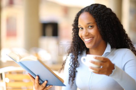 Foto de Mujer negra feliz leyendo un libro de papel tomando café en una terraza del bar - Imagen libre de derechos