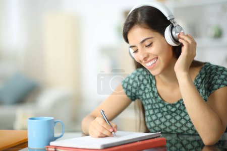 Photo for Happy student with headphone studying listening audio guide and taking notes at home - Royalty Free Image