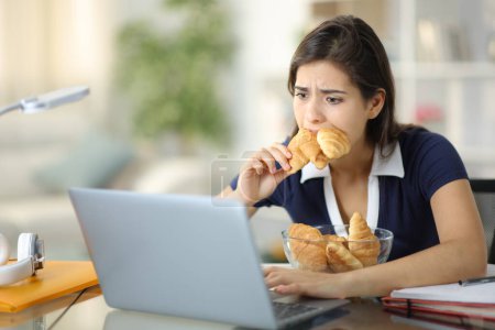 Photo for Stressed student eating a lot of bakery checking laptop at home - Royalty Free Image