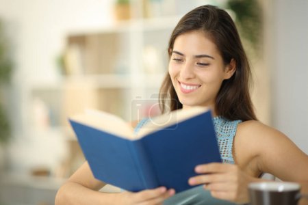 Téléchargez les photos : Femme heureuse lit livre de papier assis à la maison - en image libre de droit