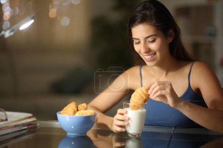 Photo for Happy woman in the night dipping bakery in milk at home - Royalty Free Image