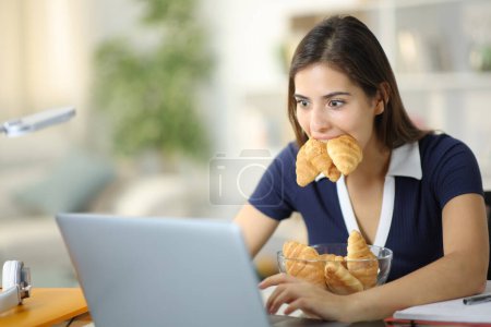 Foto de Glotón estudiante e-learning con portátil comer un montón de croissants en casa - Imagen libre de derechos