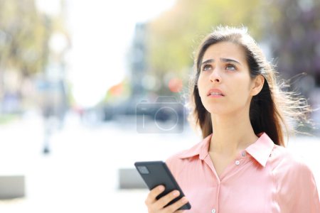 Worried woman using phone to find a place looking above standing in the street