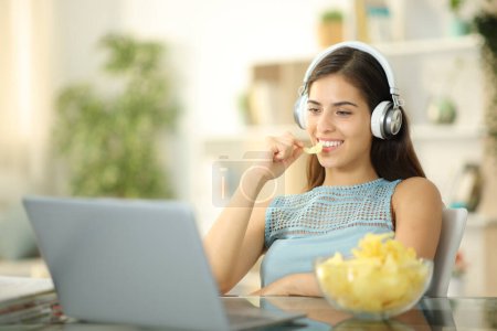 Mujer feliz comiendo bocadillo y viendo la película en el ordenador portátil en casa