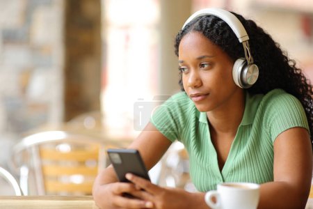 Foto de Mujer pensativa escuchando audio con teléfono y auriculares mirando hacia otro lado en una terraza del restaurante - Imagen libre de derechos