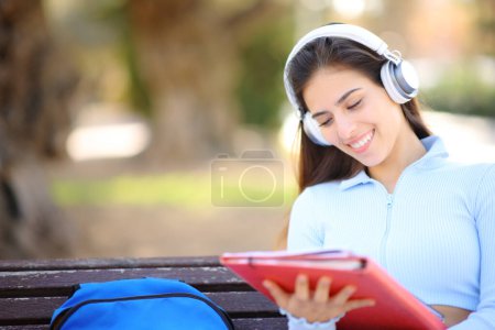 Téléchargez les photos : Étudiant heureux dans un parc écoutant un audioguide portant un casque et lisant des notes - en image libre de droit