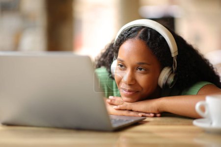 Zufriedene schwarze Frau mit Kopfhörer, die auf einer Restaurantterrasse Medieninhalte auf dem Laptop verfolgt