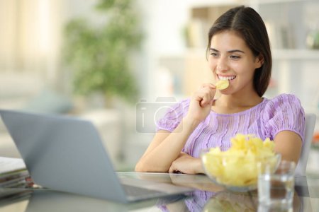 Téléchargez les photos : Femme heureuse manger des chips de pomme de terre regarder les médias sur ordinateur portable à la maison - en image libre de droit