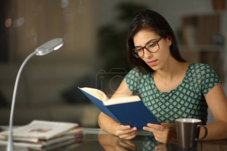 Téléchargez les photos : Femme heureuse portant des lunettes lisant un livre de papier dans la nuit à la maison - en image libre de droit