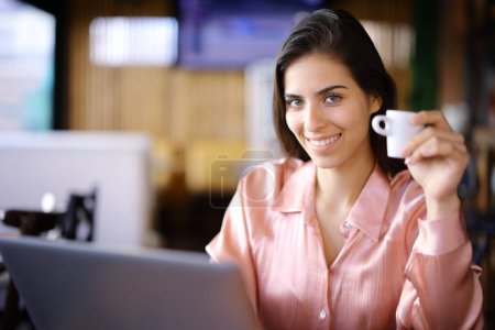 Téléchargez les photos : Heureuse femme élégante tenant une tasse de café et utilisant un ordinateur portable vous regardant dans un restaurant - en image libre de droit