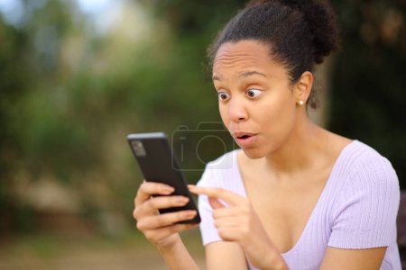 Erstaunte schwarze Frau checkt Handy draußen im Park
