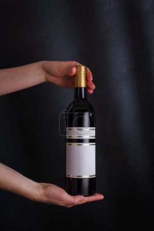 Téléchargez les photos : Mains tenant bouteille de vin rouge avec étiquette maquette, pas de marque sur fond noir. Cabernet sauvignon, merlot, pinot noir. Boisson alcoolisée, espace de copie - en image libre de droit