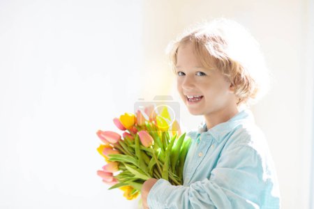 Foto de Child with flower bouquet. Mother day greeting. Little boy with bunch of tulips. Gift for mom. Spring birthday present. Kid in sunny living room holding dozens of flowers for mother. - Imagen libre de derechos
