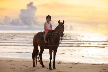 Téléchargez les photos : Enfants chevauchant à cheval sur la plage. Les enfants montent à cheval. Jolie petite fille à poney dans un ranch. Enfant et animal. Enfant en casque sûr pour la classe d'équitation. Voyagez avec un jeune enfant. Vacances au bord de l'océan. - en image libre de droit