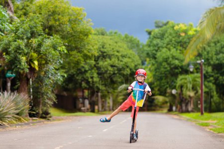 Téléchargez les photos : Petit garçon en scooter. Les enfants montent à bord. Enfant jouant dans la rue de banlieue le jour ensoleillé d'été. Casque sûr pour les enfants. Activité de plein air saine. Enfant mignon sur le chemin de l'école. - en image libre de droit