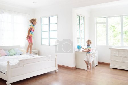 Téléchargez les photos : Enfant sautant sur le lit dans une chambre blanche avec grande fenêtre. Les enfants jouent à la maison. Saut amusant sur le lit des parents. Aménagement intérieur et chambre. Garçon à la maison. Matin ensoleillé, week-end détente en famille. - en image libre de droit