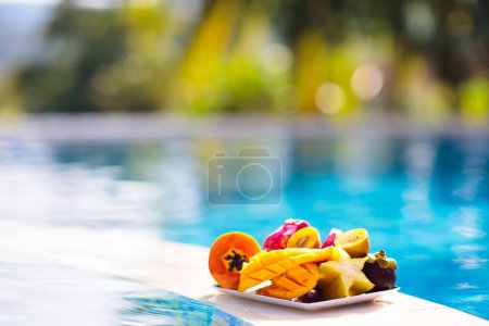 Téléchargez les photos : Assiette de fruits tropicaux à la piscine. Mangue, ananas, orange et jus frais en vacances d'été sur une île exotique. Une alimentation saine. Vacances à la plage et plaisir de nager. - en image libre de droit