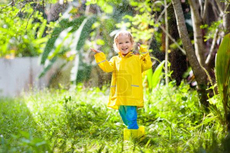 Foto de Niño jugando bajo la lluvia en el soleado día de otoño. Niño bajo ducha fuerte con paraguas de pato amarillo. Niño pequeño con zapatos impermeables de pato. Botas de goma Wellies. Otoño actividad al aire libre por tiempo lluvioso - Imagen libre de derechos