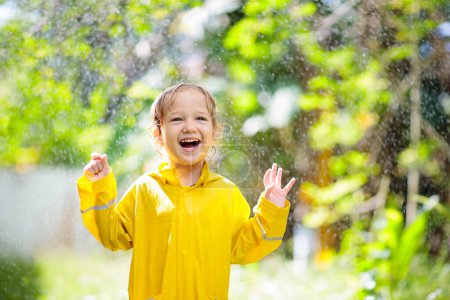 Foto de Niño jugando bajo la lluvia en el soleado día de otoño. Niño bajo ducha fuerte con paraguas de pato amarillo. Niño pequeño con zapatos impermeables de pato. Botas de goma Wellies. Otoño actividad al aire libre por tiempo lluvioso - Imagen libre de derechos