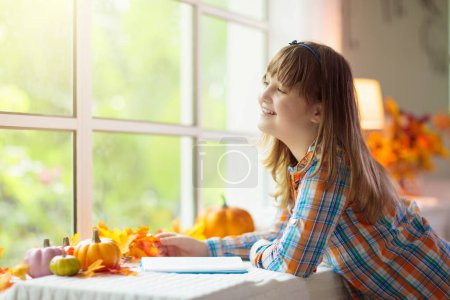 Photo for Child looking out of the window. Autumn cozy family home decoration. Kids at home in fall watching golden leaves in sunny garden. Little girl with pumpkin on Thanksgiving day or Halloween. - Royalty Free Image