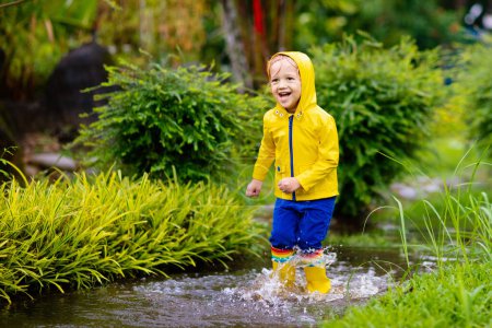 Photo for Kid playing in the rain in autumn park. Child jumping in muddy puddle on rainy fall day. Little boy in rain boots and yellow jacket outdoors in heavy shower. Kids waterproof footwear and coat. - Royalty Free Image
