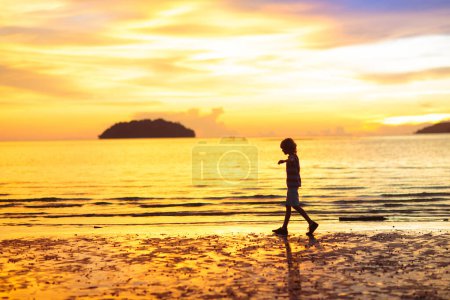 Téléchargez les photos : Enfant jouant sur la plage océanique. Il saute dans les vagues au coucher du soleil. Vacances en mer pour la famille. Petit garçon courant sur une île exotique pendant les vacances d'été. - en image libre de droit