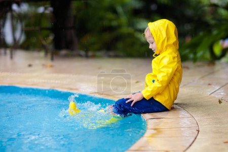 Kid jumping into swimming pool in rain. Child playing outdoors in tropical storm. Fun activities by summer rainy weather. Waterproof wear and footwear for kids. Children outdoors.