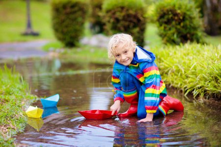 Téléchargez les photos : Enfant jouant avec bateau en papier dans la flaque d'eau. Les enfants jouent en plein air par la pluie d'automne. Automne temps pluvieux activité de plein air pour les jeunes enfants. Il saute dans des flaques boueuses. Veste et bottes imperméables pour bébé. - en image libre de droit