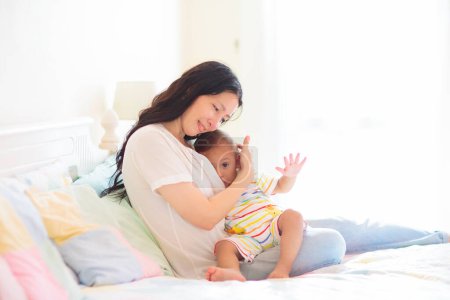 Téléchargez les photos : Young mother nursing her baby. Mom breastfeeding infant child. Asian woman and newborn boy in white bedroom with rocking chair. Nursery interior. Mother playing with laughing kid. Family at home. - en image libre de droit