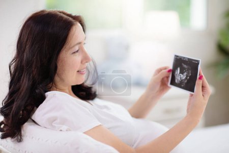 Photo for Pregnant woman at home. Young expecting mother in white bedroom. Healthy pregnancy. Female looking at baby sonogram preparing for birth. Maternity and prenatal care. - Royalty Free Image