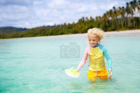 Téléchargez les photos : Enfant sur une belle plage. Petit garçon avec bateau jouet courant et sautant au bord de la mer. Vacances en mer avec un enfant. Les enfants jouent sur la plage d'été. Amusant. Les enfants nagent. Vacances en famille sur l'île tropicale. - en image libre de droit