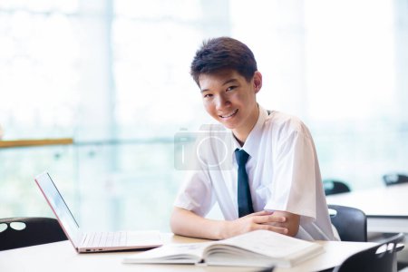 Foto de Niño asiático con portátil en la escuela. Estudiante en clase. Adolescente en uniforme haciendo deberes con la computadora en la biblioteca. Niño adolescente estudiando para la clase. Los niños estudian. Regreso a la escuela. - Imagen libre de derechos