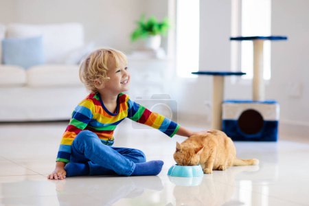 Photo for Child playing with cat at home. Kids and pets. Little boy feeding and petting cute ginger color cat. Cats tree and scratcher in living room interior. Children play and feed kitten. Home animals. - Royalty Free Image