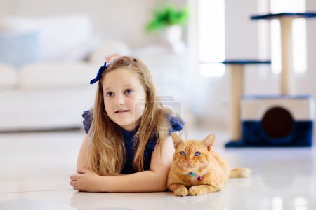 Photo for Child playing with cat at home. Kids and pets. Little girl feeding and petting cute ginger color cat. Cats tree and scratcher in living room interior. Children play and feed kitten. Home animals. - Royalty Free Image