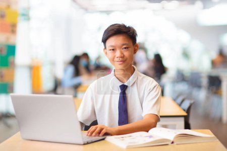 Téléchargez les photos : Asiatique garçon avec ordinateur portable à l'école. Étudiant en classe. Adolescent en uniforme faisant ses devoirs avec ordinateur à la bibliothèque. Adolescent étudiant pour la classe. Les enfants étudient. Retour à l'école. - en image libre de droit