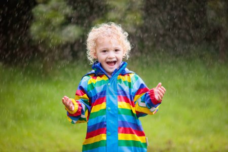 Foto de Pequeño chico rubio rizado con una chaqueta impermeable en color arco iris jugando bajo la lluvia. Niños divirtiéndose al aire libre en ducha de otoño
. - Imagen libre de derechos